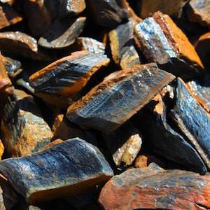 May include: A close-up of a pile of blue tiger's eye stones. The stones are rough and have a metallic sheen. They are a mix of blue, brown, and gold colors.