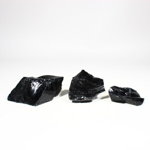 Puede incluir: Tres trozos de piedra de obsidiana negra en bruto. La obsidiana tiene una superficie brillante y reflectante y bordes irregulares y dentados. Las piedras est&aacute;n dispuestas sobre un fondo blanco, mostrando su color y textura oscuros.