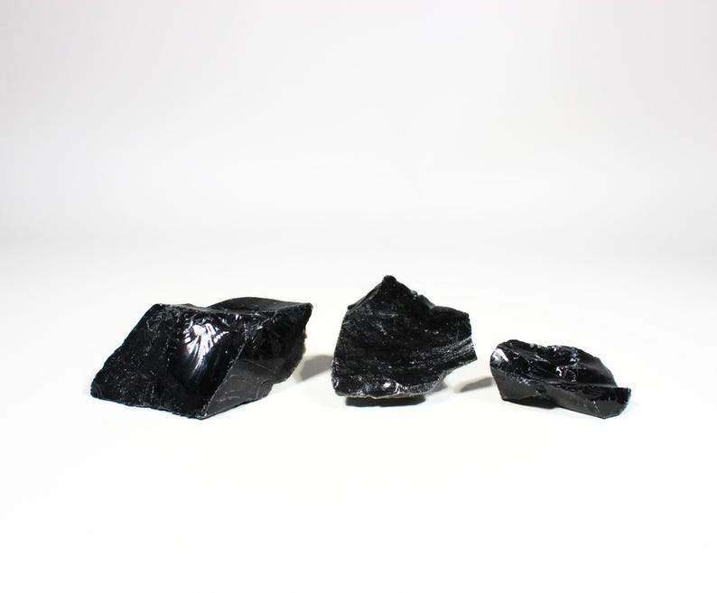 Puede incluir: Tres trozos de piedra de obsidiana negra en bruto. La obsidiana tiene una superficie brillante y reflectante y bordes irregulares y dentados. Las piedras est&aacute;n dispuestas sobre un fondo blanco, mostrando su color y textura oscuros.