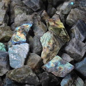 May include: A collection of rough-cut labradorite stones. The stones are primarily gray with flashes of iridescent blue, green, and gold. The natural, unpolished surfaces showcase the stone's unique textures and colors.