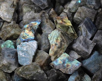 May include: A collection of rough-cut labradorite stones. The stones are primarily gray with flashes of iridescent blue, green, and gold. The natural, unpolished surfaces showcase the stone's unique textures and colors.