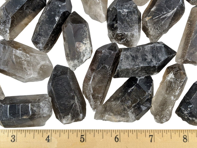 May include: A collection of smoky quartz crystal points in various sizes and shades of gray and brown. The crystals are arranged on a white surface, with a ruler providing scale. The crystals range from approximately 3 to 8 inches in length.