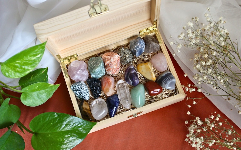 May include: A wooden box filled with a variety of colorful crystals and stones. The box is open and the crystals are arranged in a decorative pattern. The box is sitting on a red surface.