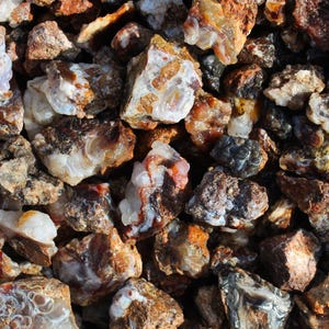 May include: A close-up view of a collection of raw, uncut gemstones. The stones vary in size and shape, displaying a range of colors including brown, orange, white, and gray. Some stones have a translucent quality, revealing internal patterns.