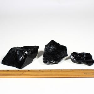 Puede incluir: Tres trozos de roca de obsidiana negra en bruto con una superficie brillante y reflectante. Se coloca una regla de madera debajo para la escala. Los trozos de obsidiana var&iacute;an en tama&ntilde;o y forma, con bordes irregulares.