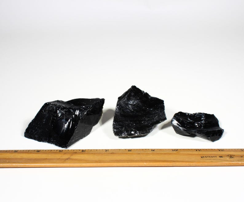 Puede incluir: Tres trozos de roca de obsidiana negra en bruto con una superficie brillante y reflectante. Se coloca una regla de madera debajo para la escala. Los trozos de obsidiana var&iacute;an en tama&ntilde;o y forma, con bordes irregulares.