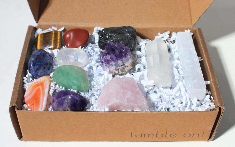 May include: A box filled with white shredded paper and 12 different tumbled crystals, including amethyst, rose quartz, clear quartz, tiger's eye, selenite, and more. The box is brown and has the words "tumble on!" printed on the bottom.