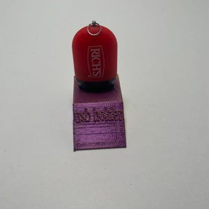 May include: Red plastic keychain with a silver ring and the word "RICH'S" printed in white on the front. The keychain is sitting on a purple 3D printed stand.