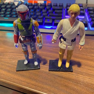 May include: Two vintage Star Wars action figures, Boba Fett and Luke Skywalker, stand on black bases. Boba Fett is wearing a green and brown suit with a helmet. Luke Skywalker is wearing a white tunic and brown pants.