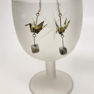 May include: Origami crane earrings with silver-tone hooks and wire. The cranes are made from patterned paper in shades of green, blue, and yellow. Each earring has a small, square, gray stone dangling below the crane.