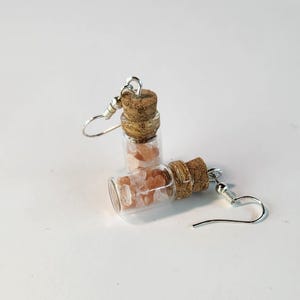 May include: A pair of earrings featuring small glass vials filled with pink crystals, each sealed with a cork stopper. The earrings have silver-colored ear wires. The background is plain white.