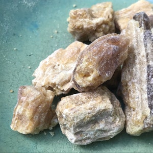 May include: Close-up of several pieces of light brown resin, some with a slightly darker brown hue. The resin is irregularly shaped and has a rough, textured surface.