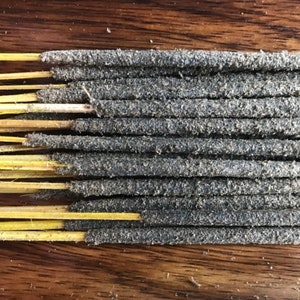 May include: Close-up of a bundle of incense sticks. Each stick has a dark gray, textured coating and a yellow wooden base. The incense sticks are arranged in parallel rows, creating a layered effect. The background is a wooden surface.