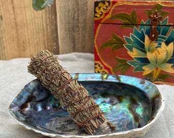 May include: A sage smudge stick rests in an iridescent abalone shell. The shell is oval-shaped with a pearlescent interior. A decorative red and gold chest is in the background. The sage bundle is tied with twine.