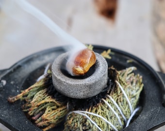 May include: A close-up of a smoldering sage bundle with a stone bowl containing a golden resin. Wisps of smoke rise from the burning sage, creating a calming and aromatic atmosphere. The sage is tied with white string.