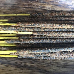 May include: Close-up of a collection of incense sticks. The sticks have a dark, textured body with a dusting of reddish-brown particles. The sticks are supported by thin, yellow stems, and are arranged on a wooden surface.