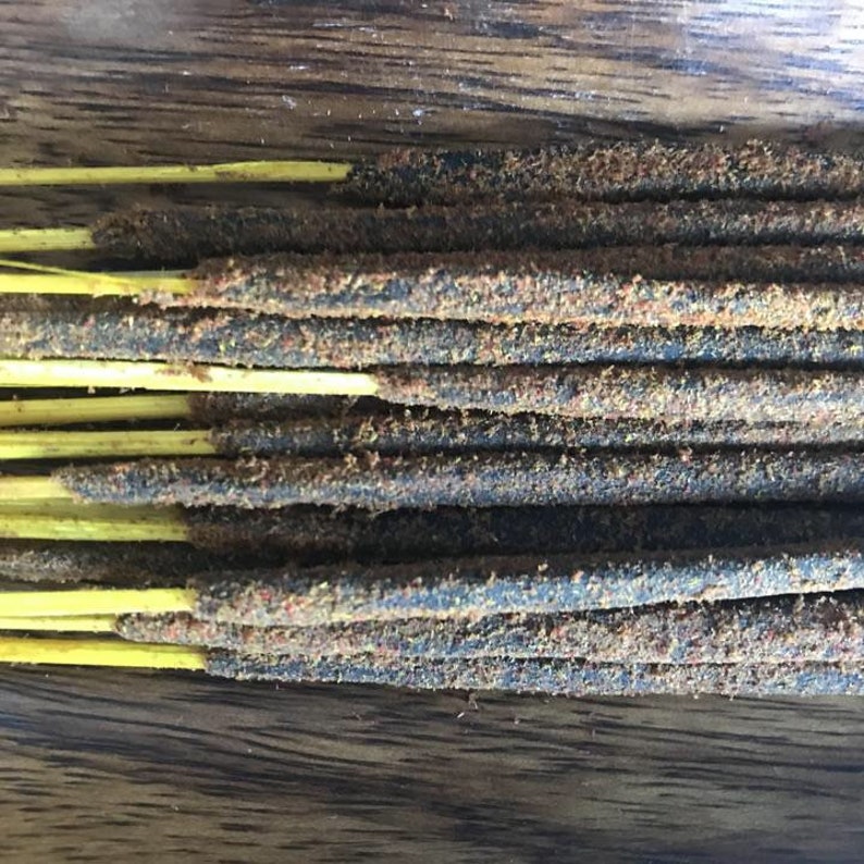 May include: Close-up of a collection of incense sticks. The sticks have a dark, textured body with a dusting of reddish-brown particles. The sticks are supported by thin, yellow stems, and are arranged on a wooden surface.