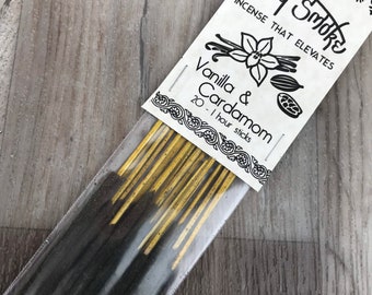 Könnte beinhalten: Eine Packung mit 20 Räucherstäbchen mit der Aufschrift "Holly Smoke Incense That Elevates Vanilla & Cardamom 20 - 1 hour sticks". Die Stäbchen sind schwarz und gold und in einer durchsichtigen Plastiktüte verpackt.