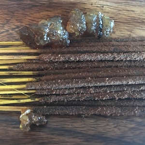 May include: A bundle of brown incense sticks with a yellow base, and several pieces of amber-colored resin on a wooden surface.