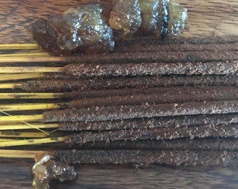 May include: A bundle of brown incense sticks with a yellow base, and several pieces of amber-colored resin on a wooden surface.