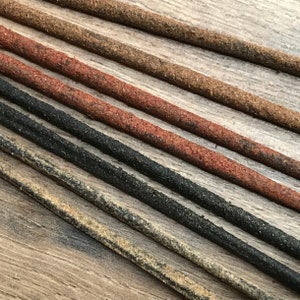 May include: Close-up of several leather cords in various colors, including brown, red, black, and tan. The cords have a textured, slightly rough surface and are arranged diagonally across a wooden surface.