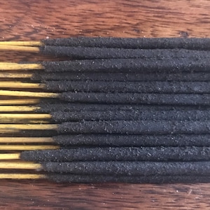 Puede incluir: Un paquete de varillas de incienso negras con puntas de madera amarilla. Las varillas están dispuestas en filas sobre una superficie de madera.