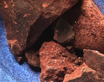 May include: Close-up of several pieces of dark red, rough-textured rock. The rocks are clustered together, with some edges catching the light. The background is a textured, deep blue surface.