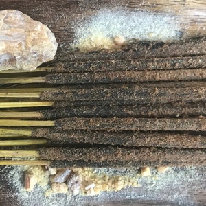 May include: A bundle of brown incense sticks with yellow bamboo bases. The sticks are arranged in a row on a wooden surface with a white and brown powder scattered around them.