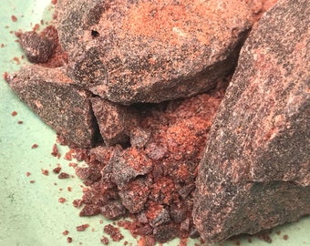 May include: Close-up of dark brown Himalayan salt crystals, some in large chunks and others in smaller pieces. The salt has a reddish hue and is scattered on a green surface.