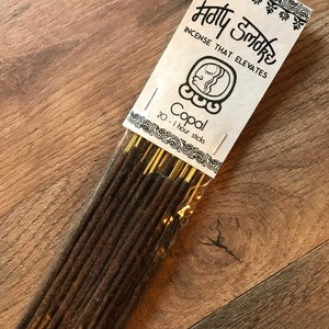 Peut inclure: Un paquet de 20 b&acirc;tonnets d'encens Copal de la marque Holly Smoke, qui se d&eacute;crit comme "l'encens qui &eacute;l&egrave;ve". Les b&acirc;tonnets sont bruns et ont une ficelle dor&eacute;e attach&eacute;e &agrave; chacun. L'emballage est blanc avec du texte noir et une illustration en noir et blanc d'une t&ecirc;te stylis&eacute;e.
