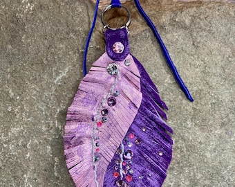 May include: A handcrafted leather feather pendant in shades of purple and pink, adorned with sparkling rhinestones and a silver-toned metal ring. The pendant is suspended from a blue cord, creating a unique and eye-catching accessory.