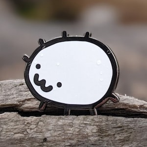 Puede incluir: Un pin de esmalte blanco con un contorno negro que representa una criatura redonda y de tipo caricatura con una cara sonriente. El pin está sentado en un trozo de madera a la deriva desgastada.