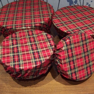 May include: Four red and gold plaid fabric bowl covers. The covers are round and have an elastic edge. The plaid pattern includes gold and black lines. The covers are on a wooden surface.