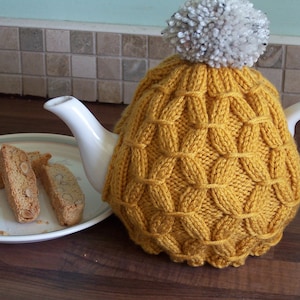 Peut inclure: Un chauffe-thé en tricot jaune avec un pompon blanc sur le dessus. Le chauffe-thé est sur une théière blanche avec une poignée et un bec verseur blancs. Il y a trois biscuits biscotti sur une assiette blanche au premier plan.