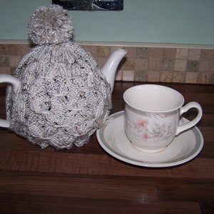 May include: A white knitted teapot cosy with a fluffy pom-pom on top. The cosy is on a teapot with a white handle and spout. A white teacup and saucer with a floral design are in the background.