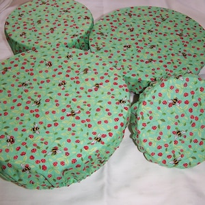 May include: Four round fabric bowl covers. They feature a pattern of bees, red berries, and green leaves on a light green background. The covers are made of a soft, flexible fabric and are designed to fit over bowls of various sizes.