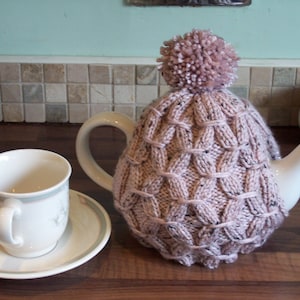 May include: A pink knitted teapot cozy with a pom-pom on top. The cozy is on a white teapot with a white teacup and saucer.