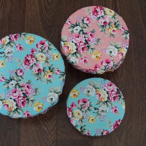 May include: Three round fabric covers with floral patterns. The largest cover is pink with white and yellow flowers. The two smaller covers are turquoise with white, pink, and yellow flowers.
