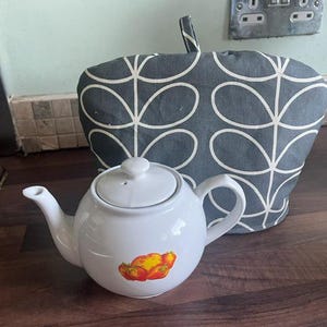 May include: A white ceramic teapot with a lid and a handle, decorated with a fruit design. A gray and white patterned tea cosy is behind the teapot. The teapot is designed for brewing tea.