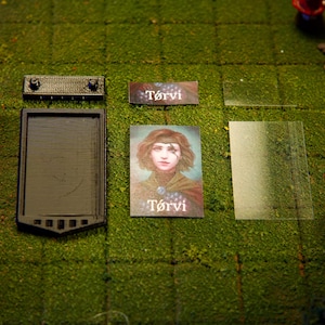 Puede incluir: Una configuraci&oacute;n de juego de mesa con una bandeja rectangular negra, una peque&ntilde;a pieza rectangular negra, una tarjeta con el nombre "T&oacute;rvi", una tarjeta de retrato de una mujer y una l&aacute;mina de pl&aacute;stico transparente. Los elementos est&aacute;n dispuestos sobre una superficie verde texturizada.