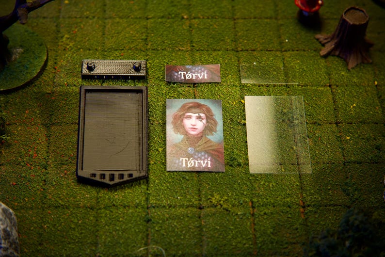 Puede incluir: Una configuraci&oacute;n de juego de mesa con una bandeja rectangular negra, una peque&ntilde;a pieza rectangular negra, una tarjeta con el nombre "T&oacute;rvi", una tarjeta de retrato de una mujer y una l&aacute;mina de pl&aacute;stico transparente. Los elementos est&aacute;n dispuestos sobre una superficie verde texturizada.