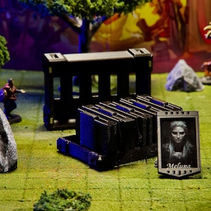 Puede incluir: Una escena de juego de mesa con figuras en miniatura, terreno y contenedores de almacenamiento negros. Un retrato enmarcado con el nombre "Melina" es visible. La escena est&aacute; ambientada sobre una superficie verde y marr&oacute;n, con &aacute;rboles y otras figuras al fondo.