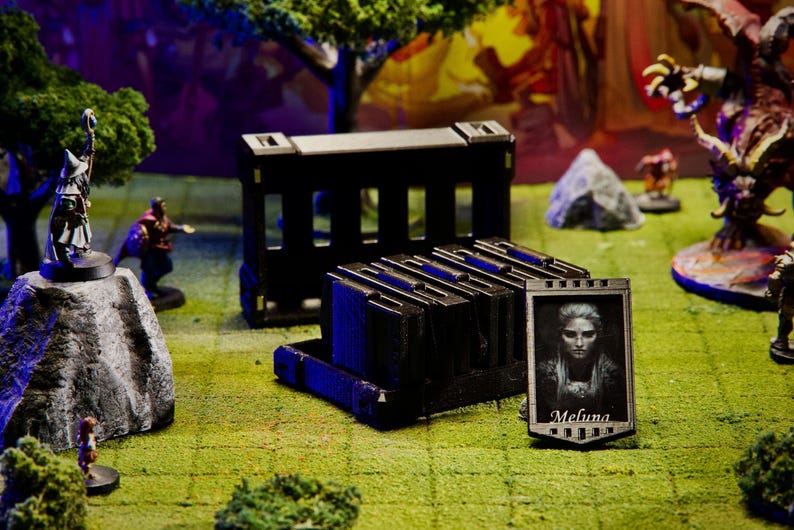 Puede incluir: Una escena de juego de mesa con figuras en miniatura, terreno y contenedores de almacenamiento negros. Un retrato enmarcado con el nombre "Melina" es visible. La escena est&aacute; ambientada sobre una superficie verde y marr&oacute;n, con &aacute;rboles y otras figuras al fondo.