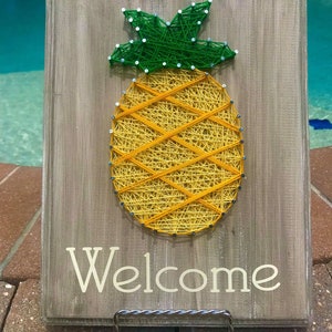 May include: A wooden sign with a string art pineapple and the word "Welcome" in white lettering.