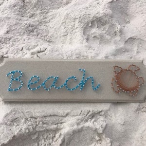 May include: A wooden sign with the word "Beach" spelled out in blue string and a brown string crab design.
