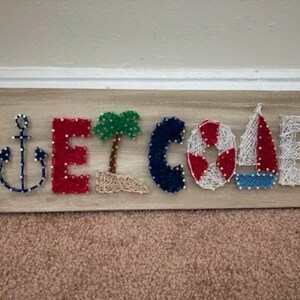 Peut inclure: Une pancarte en bois avec le mot "WELCOME" écrit en art de la ficelle. Les lettres sont décorées de thèmes nautiques, notamment une ancre, un palmier, une bouée de sauvetage et un voilier.