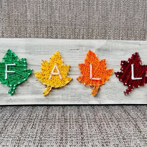 May include: A wooden sign with the word "FALL" spelled out in string art. Each letter is formed within a colorful leaf shape. The leaves are green, yellow, orange, and red.