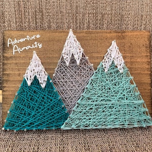 May include: String art of three mountains with white peaks. The mountains are made of teal, light blue, and gray string. The background is a wooden board with the text "Adventure Awaits" written in white.