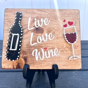 May include: Wooden sign with the words "Live Love Wine" in white lettering. The sign features a black string art wine bottle and a red string art wine glass with hearts.