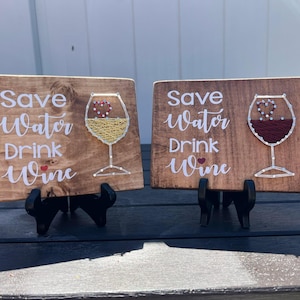 May include: Two wooden signs with the text "Save Water Drink Wine" and a string art design of a wine glass. The signs are on small black stands.